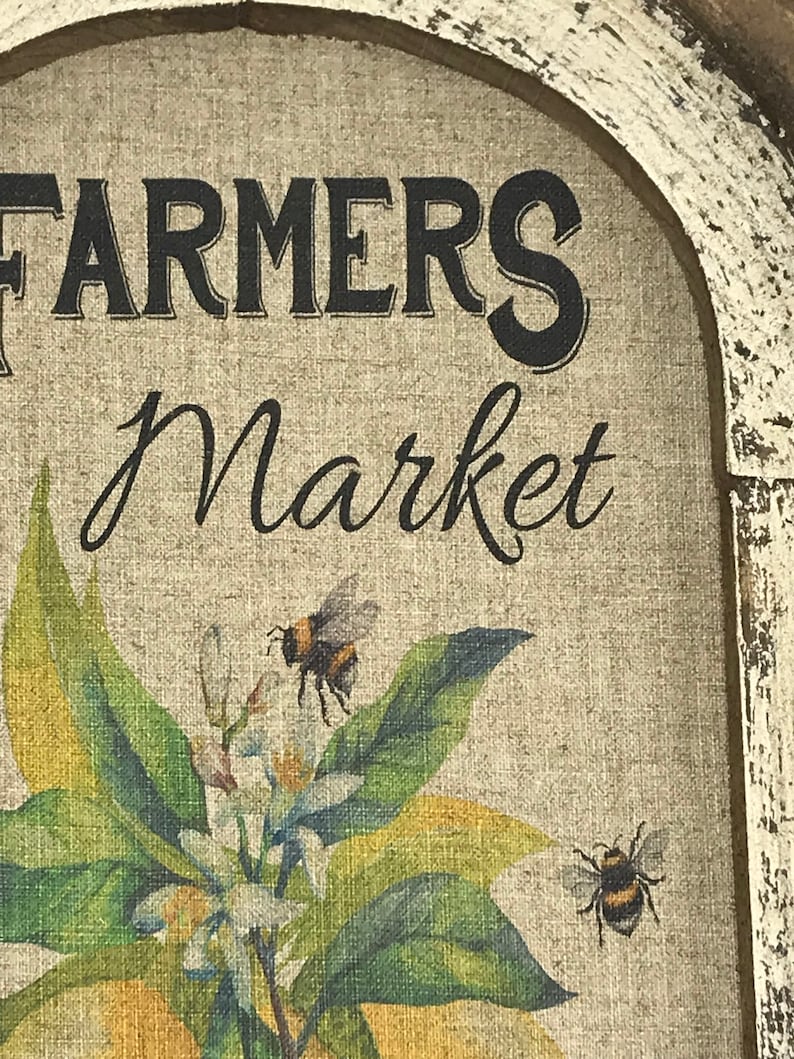 May include: A rustic wooden sign with a distressed white paint finish. The sign features the text "FARMERS Market" in black lettering and a painted image of a lemon tree with white blossoms and two bumblebees.