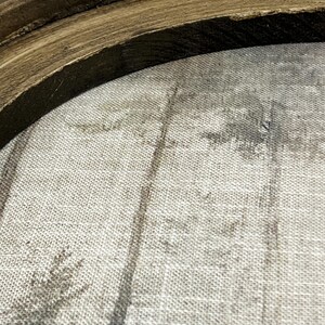 May include: Close-up of a round, framed artwork. The frame is made of dark brown, textured wood. The artwork inside features a muted, grayscale forest scene printed on a textured, light-colored fabric. The overall aesthetic is rustic and natural.