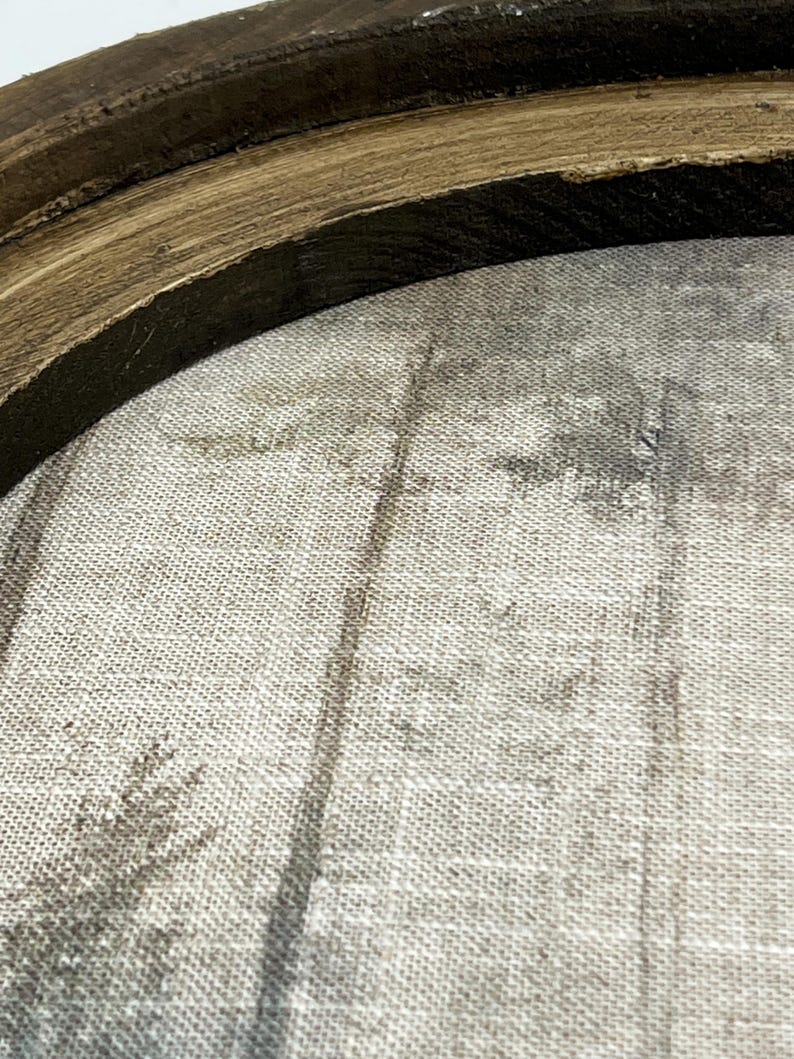 May include: Close-up of a round, framed artwork. The frame is made of dark brown, textured wood. The artwork inside features a muted, grayscale forest scene printed on a textured, light-colored fabric. The overall aesthetic is rustic and natural.
