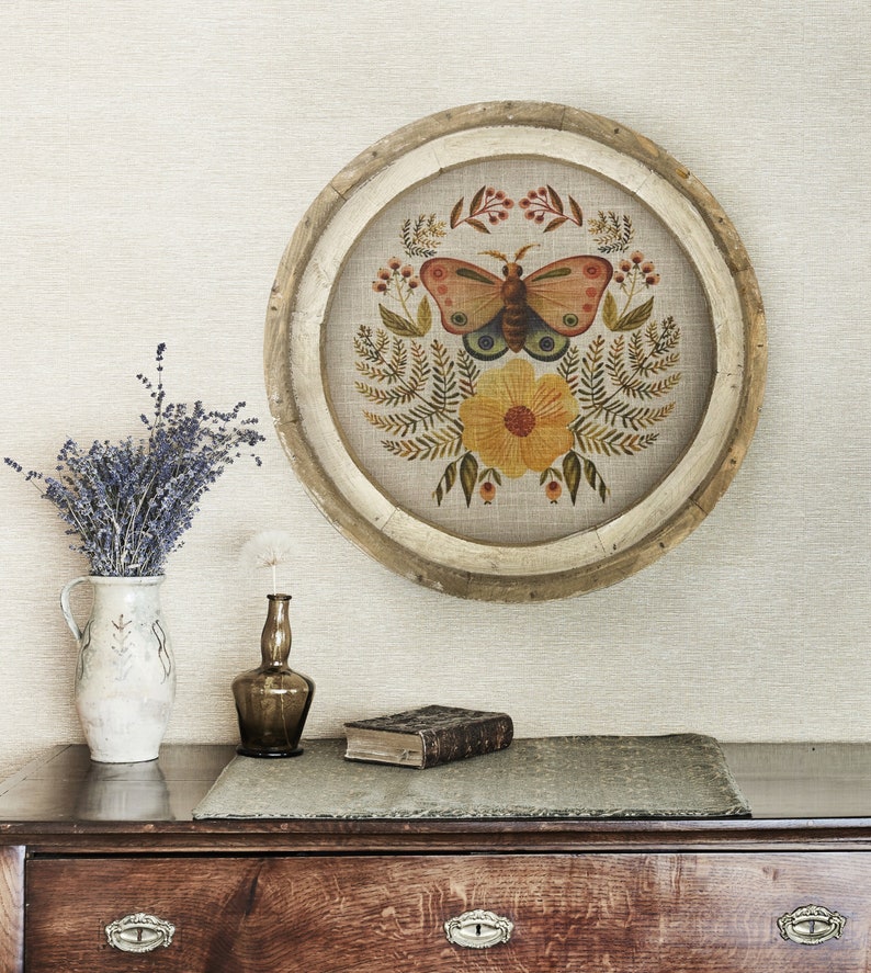 May include: A round wooden frame with a beige linen background featuring a hand-painted butterfly with orange and brown wings, a yellow flower, and green foliage.