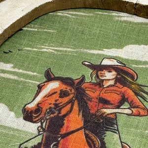 May include: A vintage-style illustration of a woman riding a brown horse. The woman is wearing a red shirt, a white cowboy hat, and blue jeans. The background is a green and white landscape.