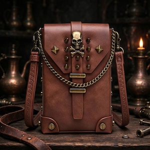 May include: A brown leather crossbody bag featuring a brass skull and crossbones emblem on the front flap. The bag is decorated with brass studs, a gold-toned chain draped across the front, and two horizontal brass bars on the vertical strap.