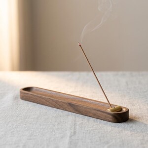 Peut inclure: Un porte-encens long et rectangulaire en bois brun foncé avec une rainure sculptée lisse. Un petit support cylindrique en laiton est placé à une extrémité, maintenant un bâton d'encens allumé avec un mince filet de fumée blanche qui s'élève.
