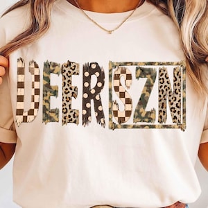 Könnte beinhalten: Ein cremefarbenes T-Shirt mit dem Schriftzug DEERSZN auf der Brust. Jeder Buchstabe zeigt ein einzigartiges Muster, darunter schwarz-weißes Schachbrettmuster, Camouflage, Leopardenmuster und schwarze Punkte auf weißem Grund.