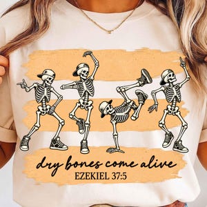 Op de afbeelding: Een crèmekleurig t-shirt met een afbeelding van vier skeletten die baseballpetten en sneakers dragen in verschillende danshoudingen. Het ontwerp bevat oranje horizontale penseelstreken en de zwarte tekst "dry bones come alive EZEKIEL 37:5".