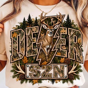 May include: A cream-coloured t-shirt featuring a graphic of a deer with large antlers centred over a lightning bolt shape. The design includes the text DEER SZN, a forest background with pine trees, autumn leaves, and pinecones in brown and green.