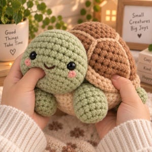 May include: A crochet turtle plush toy featuring a light green head with black safety eyes and pink stitched cheeks, a brown textured shell, and a cream-colored underside. The soft, handmade stuffed animal has a rounded shape and visible yarn stitches.