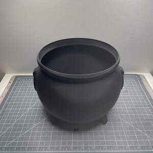 May include: A matte black cauldron with a wide circular opening, a rounded body, and three short feet. The cauldron is positioned on a gray cutting mat with white grid lines and numerical markings that show a width of approximately 10 inches.