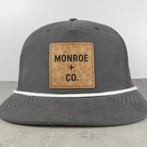 May include: A charcoal gray baseball cap featuring a square tan leather patch on the front. The patch displays a subtle floral pattern and the black text MONROE + CO. A white braided cord runs horizontally across the base of the crown.