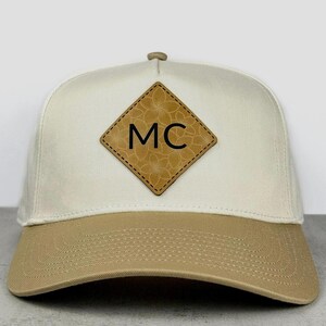 May include: A cream-coloured baseball cap with a tan brim. Centred on the front is a diamond-shaped tan patch featuring a subtle floral pattern and the black capital letters MC. The cap has a structured crown and visible stitching on the brim.