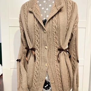 Peut inclure: Un cardigan beige en tricot torsadé avec un col, une fermeture boutonnée sur le devant et des manches longues. Le vêtement est agrémenté de petits rubans en satin marron foncé noués à la taille et sur les manches, soulignant la texture.