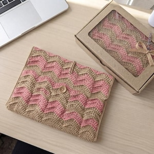May include: A rectangular crochet sleeve featuring a wavy chevron pattern in alternating tan and light pink yarn. The sleeve has a single centered wooden button closure. A matching crochet item is visible inside a brown cardboard gift box.