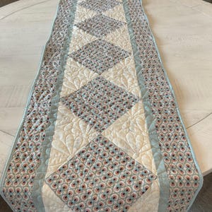 May include: A rectangular quilted table runner featuring a central row of diamond-shaped patches with a repeating circular pattern in shades of blue, pink, and cream, bordered by cream fabric with stitched floral motifs and light blue trim.