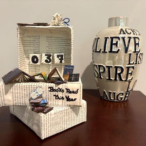May include: A decorative box covered in book pages with the text "Books Read This Year" and the number "037" displayed on small blocks. Beside it sits a white ceramic vase featuring raised dark blue lettering with words like "BELIEVE," "INSPIRE," and "LAUGH."