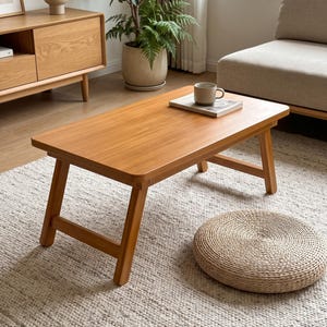 May include: A rectangular wooden coffee table with a smooth, light brown finish and four angled legs. The table features a flat surface with rounded corners and a sturdy frame, providing a stable base for items placed on top of it in a living space.
