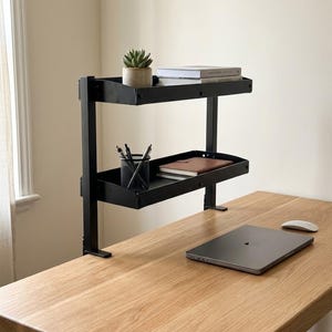 Puede incluir: Un estante de escritorio negro de metal de dos niveles con diseño de abrazadera. El estante superior sostiene una pequeña suculenta en maceta y dos libros. El estante inferior contiene un portalápices de malla metálica negra con bolígrafos.