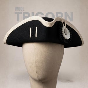 May include: A black wool tricorn hat featuring a cream-coloured trim along the upturned brim, two vertical cream-coloured stripes on the side, and a black and white pleated rosette accent with a central metallic button detail on the right side.