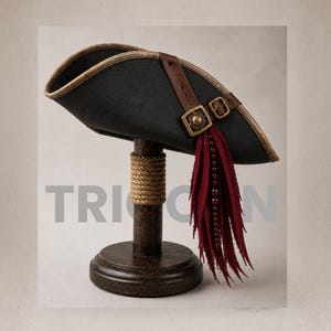 Op de afbeelding: Een zwarte driekante hoed met een bruine leren riem, een koperkleurige gesp en een decoratieve franje van donkerrode stoffen stroken en kralen. De hoed staat op een donkere houten standaard omwikkeld met touw, met het woord TRICORN op de achtergrond.