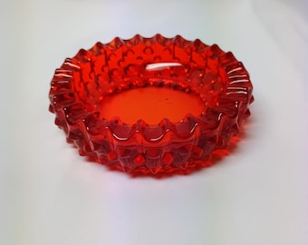 Könnte beinhalten: Ein kreisförmiger, leuchtend roter Glasaschenbecher mit einem texturierten Noppenmuster am äußeren Rand. Das Stück hat einen glatten, flachen Innenboden und ein gewelltes, gezacktes Kantendesign um den oberen Umfang, gefertigt aus durchscheinendem Glas.