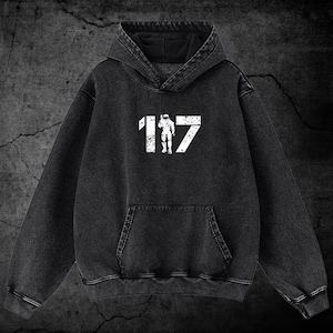 Peut inclure: Un sweat-shirt à capuche gris anthracite avec une impression graphique blanche vieillie sur la poitrine. Le motif représente le nombre 117 avec la silhouette d'un soldat debout au centre des chiffres. Le sweat possède une poche kangourou frontale.