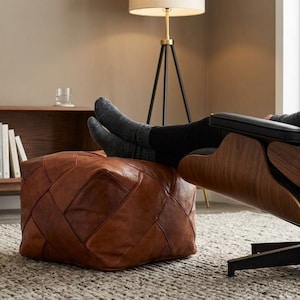 May include: A square leather ottoman featuring a patchwork design of stitched brown leather panels. The ottoman is positioned in front of a lounge chair with a dark wood shell, black leather upholstery, and a black metal swivel base.