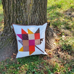 Könnte beinhalten: Ein weißes Kissen mit einem Patchwork-Ahornblatt-Design in Braun-, Orange-, Rosa- und Rottönen. Das Kissen liegt auf dem Boden vor einem Baum.