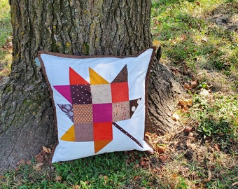 May include: A white pillow with a patchwork maple leaf design in shades of brown, orange, pink, and red. The pillow is sitting on the ground in front of a tree.
