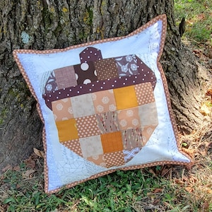 May include: A square decorative pillow with a patchwork acorn design. The acorn is made of various brown and tan fabrics, including polka dots and floral patterns. The pillow has a white border with a brown trim.