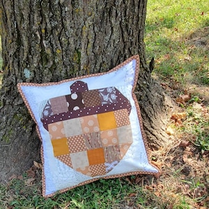 May include: A white pillow with a brown and orange patchwork acorn design. The pillow is sitting on the ground next to a tree trunk.