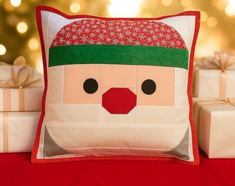 May include: A square Christmas pillow featuring a Santa Claus face design. The pillow has a red and white snowflake patterned hat, a green band, and a light pink face with black eyes and a red nose. The pillow is trimmed with a red border.