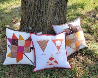 May include: Three decorative throw pillows with a fall theme. The pillows are white with brown, orange, and red patchwork designs. The pillows are labeled "pecan", "pumpkin", and "cherry".