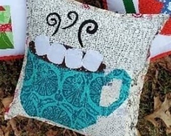 May include: A white pillow with a blue and white mug design. The mug has a brown rim and white marshmallows. The pillow has a burlap-like texture and the mug design is stitched on.