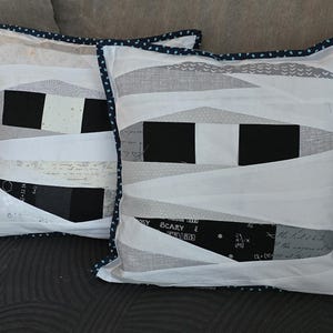 May include: Two square decorative cushions with a mummy-themed design. The cushions feature white fabric with black and grey accents, and are trimmed with a navy blue polka dot border. The design includes black rectangles and white fabric strips.