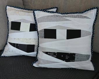 May include: Two square decorative pillows with a mummy-themed design. The pillows feature white fabric with black and gray accents, and are trimmed with a navy blue polka dot border. The design includes black rectangles and white fabric strips.