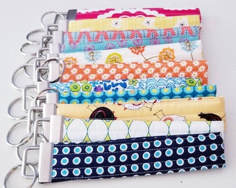 May include: A set of ten colorful fabric keychains with different patterns. Each keychain has a silver key ring attached. The keychains are made of fabric with various patterns, including polka dots, flowers, and geometric designs.