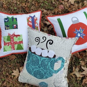 May include: Three decorative Christmas pillows with white backgrounds and red trim. One pillow features gift box designs, another has a red ornament, and the third has a mug of hot cocoa with marshmallows. The pillows are on a bed of brown leaves.