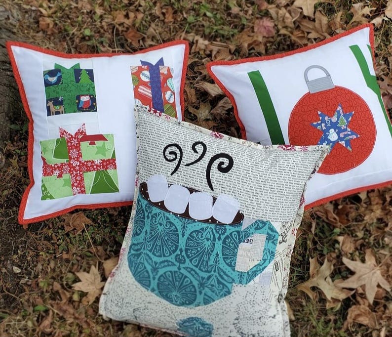 May include: Three decorative Christmas pillows with white backgrounds and red trim. One pillow features gift box designs, another has a red ornament, and the third has a mug of hot cocoa with marshmallows. The pillows are on a bed of brown leaves.