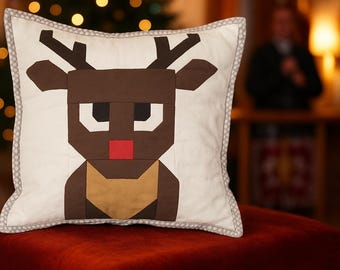 May include: Square decorative pillow with a reindeer design. The reindeer is made of brown, tan, black, red, and white fabric. The pillow has a white background with a gray dotted border. The pillow is sitting on a red surface.