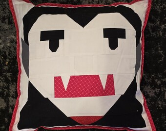 May include: Square decorative pillow with a vampire face design. The pillow features a white face with black eyes and hair, and a red mouth with fangs. The pillow is trimmed with a red border.
