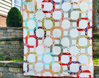 May include: A white quilt with a geometric pattern of red, green, blue, and pink fabric squares. The quilt is hanging on a stone wall.