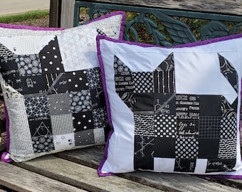 May include: Two black and white patterned throw pillows with purple trim. The pillows are shaped like a cat and feature a variety of black and white patterns, including polka dots, geometric shapes, and text.