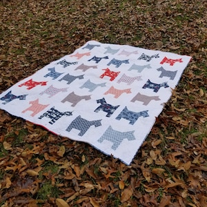 Puede incluir: Una colcha blanca con un patrón repetitivo de aplicaciones de tela en forma de perro en varios tonos de rojo, azul y gris. La colcha tiene un borde rojo fino y está confeccionada con costuras visibles en todo el fondo blanco.