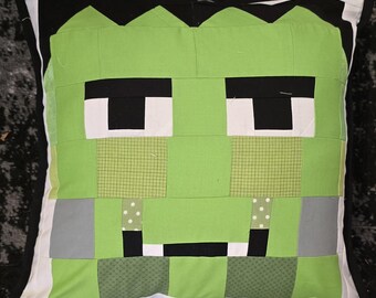 May include: Square decorative pillow with a patchwork design featuring a green monster face. The pillow has a black, white, and green color scheme, with a black border and white backing. The monster face has black eyes and a stitched mouth.