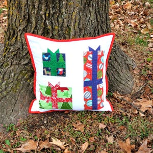 May include: A white Christmas-themed pillow with a red border, featuring three gift box designs. The gift boxes are green and red with blue ribbon accents. One box has a bird, deer, and owl design.