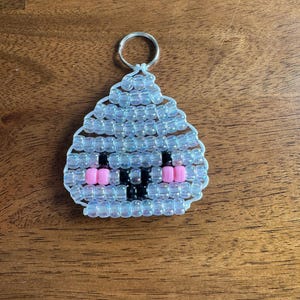 May include: A keychain crafted from iridescent clear plastic pony beads arranged in a triangular shape. The design features a face created with black beads for eyes and a nose, and pink beads for cheeks, all strung on white cord with a metal ring.