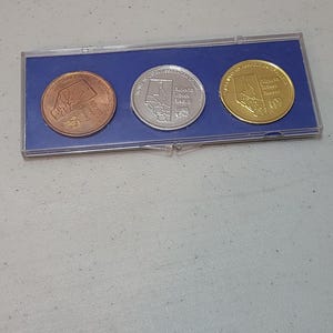 May include: A rectangular clear plastic display case containing three circular commemorative coins in copper, silver, and gold tones. Each coin features a map of Alberta and the text Alberta Travel Award, set against a blue velvet-like insert.