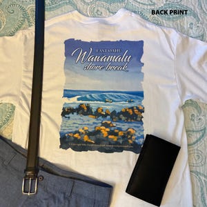 May include: A white t-shirt featuring a back print of a coastal scene with the text "EAST OAHU Wawamalu shore break" above a blue ocean wave and dark rocks. The shirt is displayed alongside a black leather belt and a black rectangular wallet.