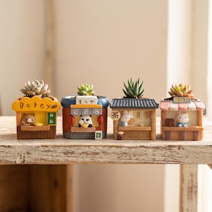 May include: Four miniature resin planters shaped like small shop stalls, each containing a small succulent. The stalls feature a yellow bakery, a blue panda noodle shop, a gray tea house with a rabbit, and a pink cat-themed shop.