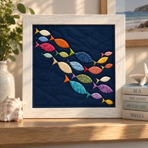 Op de afbeelding: Een vierkant wandkunstwerk met een donkerblauwe gewatteerde stoffen achtergrond en een school kleine, veelkleurige stoffen vissen in tinten oranje, geel, groen, blauw, paars en roze, gevat in een lijst met een lichte houtnerf.