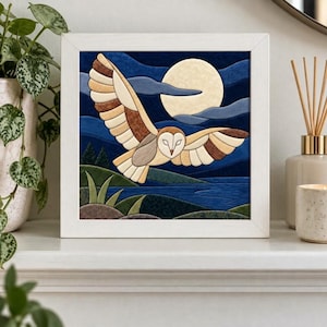 May include: A square framed fabric art piece depicting a barn owl in flight against a night sky with a large pale moon. The scene includes dark blue clouds, a body of water, green hills, and dark trees, all rendered in layered fabric textures.